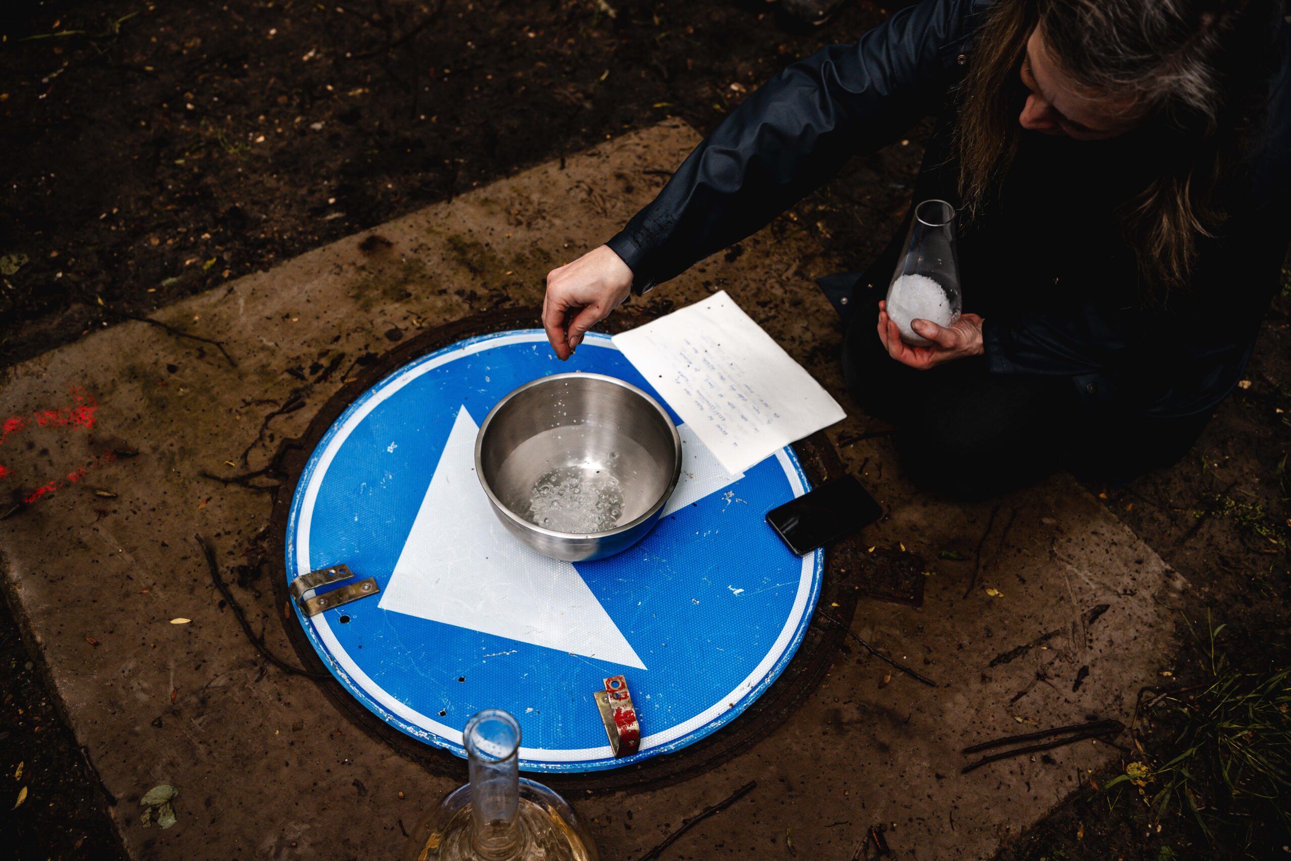 picture of a woman putting salt into an inox bowl placed on a blue and white traffic sign that is lying on the ground